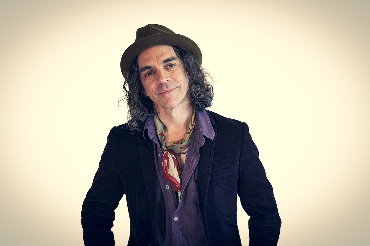 Australian blues musician Nathan Cavaleri smiling in a fedora, dark jacket, purple shirt, and patterned neck scarf against a plain backdrop.